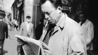 L'image représente un homme dans une rue animée, semblant lire un journal avec attention. Il porte un manteau clair et une cigarette à la bouche. En arrière-plan, on peut voir d'autres passants et des commerces, ce qui donne une ambiance des années passées. La scène évoque une atmosphère de réflexion et d'intérêt pour l'actualité.