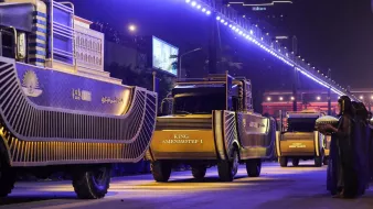 L'image montre un défilé nocturne avec des camions décorés, probablement pour une célébration ou un événement spécial. Les camions portent des inscriptions, dont "KING AMENHOTEP I", et sont éclairés par des lumières vives. En arrière-plan, on aperçoit un bâtiment, peut-être un hôtel, et des foules observant l'événement. Un personnage en costume traditionnel est également visible, ajoutant à l'atmosphère festive et culturelle.