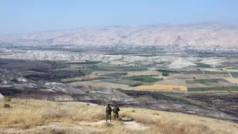 L'image montre un paysage montagneux et rural. Au premier plan, deux personnes se tiennent de dos, probablement des soldats ou des randonneurs, observant la vallée en contrebas. La vallée est composée de champs et de terrains cultivés, avec des couleurs variées allant du vert au jaune. À l'arrière-plan, des collines et des montagnes se profilent sous un ciel bleu clair. L'ensemble dégage une impression de tranquillité et de vaste espace naturel.