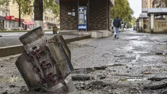 L'image montre un paysage urbain marqué par la destruction. Au premier plan, on voit un obus ou un engin explosif détérioré, partiellement enfoui dans le sol. Le bitume est endommagé autour de l'obus, laissant des fissures et des débris. En arrière-plan, un homme marche sur le trottoir, près d'un bâtiment qui semble intact. L'atmosphère est sombre et témoigne des conséquences d'un conflit ou d'un événement violent. Les arbres sur le bord de la route ajoutent une touche de verdure, mais le contexte reste très préoccupant.