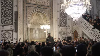 در تصویر، گروهی از افراد در یک مکان مذهبی یا تاریخی تجمع کرده‌اند. در وسط تصویر، فردی با لباس نظامی در حال سخنرانی یا بیان مطلبی به جمعیت است. نفرات در اطراف او با تلفن‌های همراه خود مشغول فیلم‌برداری یا عکاسی هستند. محیط اطراف آن‌ها تزئینات زیبا و نورپردازی چشمگیری دارد، همچنین یک چراغ آویز بزرگ در بالای سر گروه دیده می‌شود که به فضا زیبایی می‌بخشد. دیوارها با نقوش و طراحی‌های پیچیده تزئین شده‌اند و جو عمومی تصویر نشان‌دهنده شور و شوق جمعیت است.