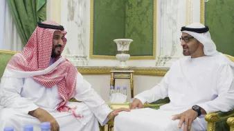 L'image montre deux hommes en vêtements traditionnels. L'un porte une robe blanche avec un manteau rouge et blanc, tandis que l'autre est habillé en blanc avec un couvre-chef noir. Ils sont assis côte à côte sur des fauteuils dans un cadre élégant, décoré de vert et d'or. Les deux hommes semblent engagés dans une conversation amicale. Sur la table devant eux, on aperçoit des bouteilles d'eau.