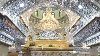 الصورة تظهر مشهداً داخلياً في مكان ديني، حيث تتواجد الكثير من النساء المحجبات داخل قاعة مزينة بتفاصيل معمارية جميلة ومزيّنة بالزخارف والجدران المغطاة بالزجاج اللامع. يتوسط القاعة مصباح ضخم معلق، ويبدو أن هناك ضريحاً أو مكان مقدس في الخلفية مزين بالعناصر الذهبية والكتابات الإسلامية. الأجواء تعكس روحانيّة وعمق الإيمان، حيث يتجمع الناس في مكان يتسم بالهدوء والتأمل.