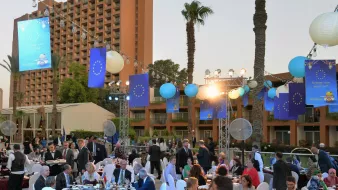 L'immagine mostra un evento elegante all'aperto, probabilmente un ricevimento o una cena di gala. Ci sono diverse tavole decorate con tovaglie scure e sedie bianche, alcune delle quali sono occupate da ospiti in abbigliamento formale. Si possono vedere lampade cinesi appese e bandiere che rappresentano l'Unione Europea. Sullo sfondo ci sono palme e un edificio, mentre la luce del tramonto crea un'atmosfera suggestiva. Le persone sembrano conversare e godere della serata.