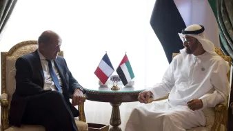 The image shows two men seated at a table in a luxurious setting. They are engaged in conversation, appearing to be in a diplomatic or formal meeting. There are flags on the table representing France and the United Arab Emirates. The background features elegant curtains and decor. One man is dressed in a suit, while the other is wearing traditional Emirati attire.