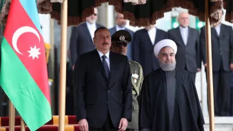 این تصویر دیداری رسمی را نشان می‌دهد که در آن دو فرد به همراه یکدیگر ایستاده‌اند. یکی از آنها در لباس رسمی و دیگری در لباس روحانی به چشم می‌خورد. در پس‌زمینه پرچم آذربایجان دیده می‌شود و تعدادی از افراد دیگر نیز در پس‌زمینه حاضرند. فضای کلی تصویری رسمی و دولتی به نظر می‌رسد.