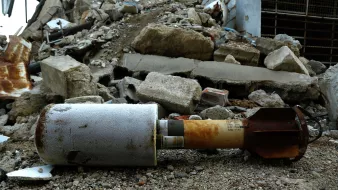 L'image montre un obus ou une roquette qui est reposé sur un sol poussiéreux, entouré de débris et de morceaux de béton. En arrière-plan, on peut voir un paysage marqué par des ruines, suggérant une scène de destruction. L'objet semble endommagé et perdu au milieu de l'effondrement des structures environnantes.