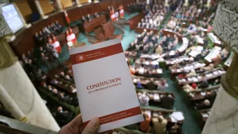 The image shows a parliamentary session in Tunisia, with a focus on a hand holding a book titled "Constitution de la République Tunisienne" (Constitution of the Tunisian Republic). In the background, a large assembly of representatives is seated in a formal, legislative setting, likely discussing or voting on matters related to the constitution or other important legislative issues. The architecture of the chamber features ornate details, and national flags are visible, emphasizing the official nature of the event.