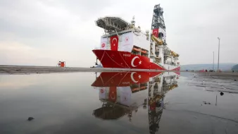 L'image montre un navire de forage maritime, nommé "Yavuz", qui est amarré au port. Le bateau arbore des couleurs dominantes de rouge et blanc, représentant le drapeau turc. On peut également voir des équipements de forage sur le pont du navire. Dans la partie inférieure de l'image, il y a des réflexions du navire dans une flaque d'eau sur le sol. Le ciel en arrière-plan est nuageux, créant une atmosphère calme.
