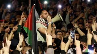 L'image montre un grand rassemblement de personnes, probablement lors d'une manifestation. Au premier plan, un homme utilisant un mégaphone pour parler à la foule. Il est entouré de nombreux participants qui lèvent les bras et brandissent des drapeaux, notamment des drapeaux palestiniens. L'atmosphère semble électrisante et engagée, avec des gens qui filment l'événement avec leurs téléphones. La scène dénote une forte mobilisation et un sentiment collectif.