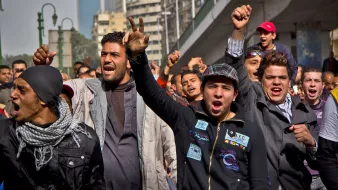 L'image montre un groupe de jeunes manifestants dans une rue animée. Ils expriment des émotions fortes, avec des visages déterminés et des gestes de protestation. Certains lèvent les bras et serrent les poings, tandis que d'autres crient. L'arrière-plan présente des bâtiments urbains, et l'atmosphère générale semble être celle d'une mobilisation collective. Les personnes sont habillées de manière variée, reflétant un mélange de styles.
