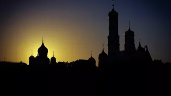 تظهر في الصورة silhouettes لعدد من الكنائس مع قبابها وأبراجها في لحظة غروب الشمس. الخلفية تميل إلى الألوان الدافئة للغروب، مع تدرجات من الأصفر إلى الأزرق الداكن. تبرز المعالم المعمارية بشكل جميل ضد السماء، مما يضفي على الصورة جوًا من الهدوء والجمال الروحي.