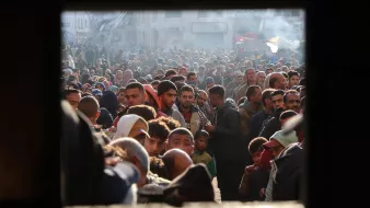 L'immagine ritrae una grande folla di persone in un'area urbana. Si osservano uomini, donne e bambini, tutti concentrati in uno spazio ristretto. L'atmosfera sembra intensa e caotica, con molte persone che si affollano. La luce del sole filtra tra gli edifici, creando un contrasto tra ombre e zone illuminate. La scena potrebbe suggerire un evento significativo o una situazione di emergenza.