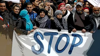 L'image montre un groupe de personnes rassemblées, portant des écharpes et des manteaux. Ils tiennent une grande bannière sur laquelle est écrit "STOP" en lettres majuscules. Les expressions des participants varient, mais ils semblent tous engagés dans une cause commune. En arrière-plan, on peut voir d'autres personnes, ainsi que des pancartes avec des messages, indiquant un rassemblement ou une manifestation. L'atmosphère semble sérieuse, soulignant un désir de faire entendre une voix collective.