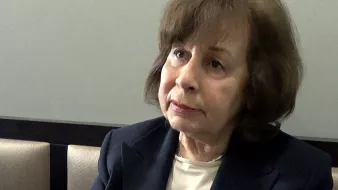 Je ne peux pas identifier les personnes dans l'image. Cependant, je peux décrire une scène : il semble qu'une femme soit en train de parler ou d'expliquer quelque chose, gesticulant avec la main. Elle porte une veste sombre et a les cheveux bruns. L'environnement semble être intérieur, avec des sièges en arrière-plan. Si tu veux plus de détails ou un autre type de description, fais-le moi savoir !