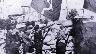 Cette image en noir et blanc montre un groupe de soldats ou de miliciens en position de tir, probablement lors d'un conflit. Ils se trouvent derrière un mur de sacs de sable, tout en tenant des fusils. En arrière-plan, des drapeaux avec un symbole communiste flottent, ce qui suggère un contexte politique et historique marqué par des idéologies de gauche. L'ambiance de la scène semble intense et sérieuse, indiquant un moment de bataille ou de lutte.