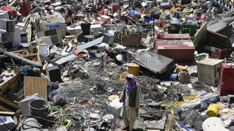 The image shows a cluttered scene filled with a massive amount of discarded items, including various types of debris, appliances, and objects that appear to be from different categories. Amidst this chaotic environment, a person is seen walking through the debris, navigating the piles of junk. The area looks like a dump or recycling site, emphasizing the vast accumulation of waste and the struggle to find salvageable materials within.