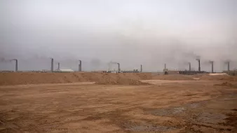 این تصویر یک محیط صنعتی را نشان می‌دهد که در آن دود از دودکش‌های بلند بیرون می‌آید. آسمان به طور کلی ابری و خاکستری به نظر می‌رسد و هوای منطقه آلوده است. زمین خاکی و خشک به نظر می‌رسد و هیچ گونه پوشش گیاهی قابل مشاهده نیست. این صحنه می‌تواند نشان‌گر فعالیت‌های صنعتی و مشکلات زیست‌محیطی باشد.