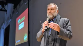 Nell'immagine si vede un uomo, probabilmente un relatore, che sta parlando al pubblico. Indossa un abito elegante e ha una sciarpa scura. Tiene un microfono in mano e sembra essere coinvolto in un discorso. Sullo sfondo c'è un grande schermo con delle grafiche colorate, ma il contenuto di ciò che viene mostrato non è visibile. L'atmosfera dell'immagine sembra essere formale e interattiva.