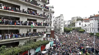 L'image montre une grande foule rassemblée devant un bâtiment, manifestant dans une ville. De nombreuses personnes sont visibles sur les balcons du bâtiment, brandissant des drapeaux et exprimant leur soutien à la manifestation. La rue est remplie de manifestants, et l'atmosphère semble vibrante et dynamique, avec un mélange de drapeaux et de pancartes. Le paysage urbain environnant, composé de divers bâtiments, ajoute à l'environnement de la scène.