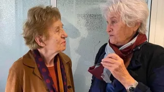 L'image montre deux femmes âgées engagées dans une conversation. Elles semblent amicales et souriantes. L'une porte une veste marron avec un foulard à pois, tandis que l'autre est habillée de manière similaire, avec une écharpe. L'arrière-plan est flou, mettant en valeur leur interaction.