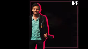 L'image montre un homme souriant, portant une chemise turquoise sous un veston noir. Il a une carte d'identification accrochée à son cou et tient un appareil photo à la main. Le fond est principalement noir, avec un contour rose qui souligne la silhouette de l'homme. En haut à droite, le logo "RSF" est visible.