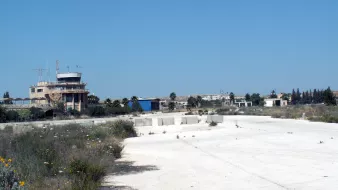La imagen muestra un paisaje de un área desierta, posiblemente un antiguo aeropuerto o base aérea. En el fondo se observa una torre de control o edificio administrativo, rodeado de vegetación y estructuras en desuso. El suelo es de color claro y parece estar cubierto de hierbas y flores silvestres, lo que sugiere abandono. El cielo es de un azul despejado, lo que le da un contraste al paisaje.