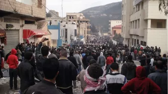 L'image montre une grande foule de personnes rassemblées dans une rue, entourée de bâtiments. Les gens semblent très nombreux, et certains portent des vêtements sombres ou des écharpes. L'atmosphère paraît tendue ou mobilisée, avec des manifestations potentielles. En arrière-plan, on aperçoit des constructions et des collines. L'ensemble donne une impression de mouvement collectif et d'unité.