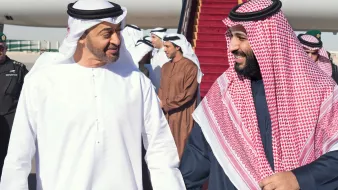 L'image montre deux hommes vêtus de vêtements traditionnels arabes, marchant côte à côte sur un tapis rouge. Ils semblent échanger un moment amical, souriant l'un à l'autre. En arrière-plan, plusieurs autres personnes sont présentes, ainsi qu'un avion stationné. L'atmosphère semble chaleureuse et détendue.