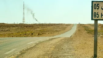 L'image montre un paysage désertique avec une route qui s'étend à perte de vue. À droite, un panneau indiquant "Hassi R'Mel" avec des chiffres. En arrière-plan, on aperçoit une tour de communication et une colonne de fumée émanant d'une installation, probablement liée à l'industrie pétrolière. Le sol est aride, et il n'y a pas beaucoup de végétation environnante, illustrant un environnement rural et isolé.