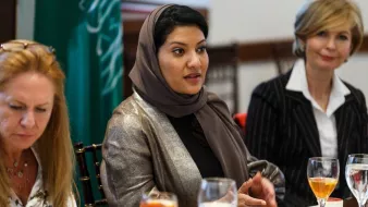 L'image montre un groupe de femmes assises autour d'une table, en train de discuter. Une femme au centre porte un hijab et un vêtement aux couleurs neutres, semblant engagée dans la conversation. À sa gauche, une autre femme aux cheveux longs et blonds, légèrement penchée, écoute attentivement. À droite, une femme en tailleur noir et blanc observe également. Sur la table, on peut voir des verres de boisson et de la vaisselle, avec des éléments de nourriture. L'environnement semble formel, propice à une réunion ou un déjeuner d'affaires.