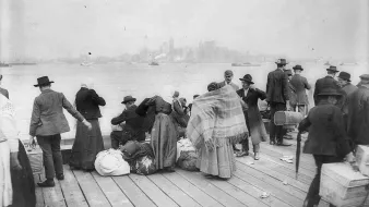 L'image montre un groupe de personnes rassemblées sur un quai, probablement à une époque antérieure. On peut voir des individus portant des habits typiques de l'époque, dont des chapeaux, et certains sont emmitouflés dans des couvertures. Ils semblent s'affairer autour de bagages et de marchandises, évoquant des thèmes de migration ou de transit. En arrière-plan, on aperçoit des bâtiments ainsi que des bateaux sur l'eau, suggérant un port animé. L'atmosphère semble à la fois chargée et historique.