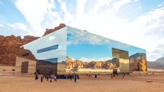 L'image montre un bâtiment moderne et futuriste situé dans un paysage désertique. La structure est entièrement recouverte de miroirs, reflétant le ciel et les montagnes environnantes, ce qui lui permet de se fondre harmoniellement dans le paysage. Au premier plan, un groupe de personnes, vraisemblablement des visiteurs, se regroupe sur le sable. L'atmosphère est paisible, et le ciel est parsemé de nuages, créant un contraste avec les teintes désertiques. L'architecture innovante et les réflexions ajoutent une dimension visuelle fascinante à l'environnement naturel.
