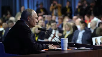 L'image montre un homme assis à une table de conférence, parlant peut-être lors d'une conférence de presse ou d'un discours. Il semble concentré et fait face à un public, qui est partiellement visible en arrière-plan. On aperçoit des journalistes prenant des notes ou photographiant. Des micros sont placés devant lui, et une tasse blanche est posée sur la table. L'atmosphère paraît sérieuse et attentive.