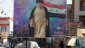 الصورة تظهر لوحة إعلانات كبيرة تتضمن صورة لشخص يرتدي زيًا دينيًا يتوسط خلفية من الجماهير. يبدو أنه يحمل علمًا، ويتقدم في مشهد يعكس حماسًا واحتشادًا. في أسفل اللوحة، هناك كتابة تشير إلى هوية شخصية أو جهة. في المقدمة، يظهر رجل يسير بجوار اللوحة، مما يعكس الحياة اليومية في المكان. الصورة تنقل أجواء من النشاط السياسي والتفاعل الاجتماعي.