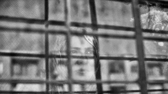 L'image montre une femme qui apparaît derrière une grille. Le contraste entre le flou et les barreaux de la grille donne une impression de séparation ou d'enfermement. La femme semble contemplative, et son expression peut évoquer des émotions profondes. Les nuances de noir et blanc ajoutent une atmosphère mélancolique à la scène.