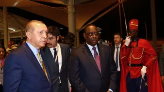 L'image montre deux hommes en costume, marchant ensemble dans un cadre officiel. L'un d'eux semble être un dirigeant turc, tandis que l'autre représente le Sénégal. Ils sont entourés de plusieurs personnes en arrière-plan, et un garde d'honneur en uniforme rouge se tient à proximité. L'atmosphère paraît sérieuse et formelle, typique d'une rencontre diplomatique.