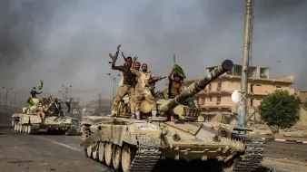 L'image montre des tanks avancant sur une route dans un environnement urbain. Plusieurs soldats sont visibles sur les tanks, certains levant les bras en signe de victoire ou de célébration. L'arrière-plan présente des bâtiments endommagés et une atmosphère chargée de fumée, ce qui suggère un contexte de conflit. Les couleurs sont principalement terreuses, avec des teintes de vert et de beige des tanks et des uniformes des soldats.
