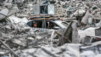 L'image montre un paysage dévasté avec des décombres sur le sol. Au centre, on aperçoit une voiture dans laquelle se trouvent deux personnes. La voiture semble être coincée parmi les débris, qui comprennent des morceaux de béton et d'autres matériaux d'une structure effondrée. L'ambiance générale dégage une impression de destruction et de chaos, témoignant d'une situation difficile.