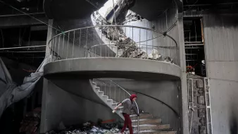 L'immagine mostra una scala a chiocciola danneggiata all'interno di un edificio in rovina. La struttura presenta macerie sparse e parte del soffitto è crollato, creando un'apertura sopra la scala. Una persona, vestita con un abbigliamento rosso e coperta da un copricapo, sta camminando sulla scala tra le macerie. L'atmosfera è quella di un luogo in crisi, con segni evidenti di distruzione e abbandono.