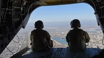 The image shows two military personnel seated inside a helicopter, looking out through the open door. They're wearing tactical gear and helmets, and the view below features a landscape with land and water. Various structures can be seen in the distance, highlighting a bustling environment beneath them. The scene conveys a sense of operation and readiness, capturing the perspective of those in flight.