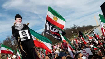 L'immagine ritrae una manifestazione in cui si vedono molte persone riunite, molte delle quali tengono in mano bandiere dell'Iran. C'è un grande cartello con la scritta "Woman, Life, Freedom" e un uomo che tiene una sagoma con la figura di un anziano, che sembra simboleggiare una figura autoritaria. Sullo sfondo si possono notare manifestanti con vari cartelli e bandiere, tutti uniti per esprimere il loro messaggio di protesta. La scena trasmette un forte senso di unità e determinazione tra i partecipanti.