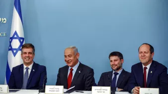 L'image montre un groupe de quatre hommes assis à une table, souriant et discutant. En arrière-plan, il y a un drapeau israélien. Les hommes portent des costumes et des cravates, et il semble qu'ils soient en train de participer à une conférence de presse ou à un événement officiel. L'ambiance est décontractée mais professionnelle.