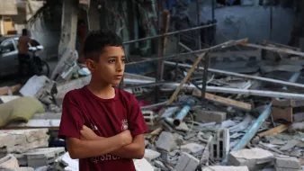 L'immagine mostra un ragazzo con le braccia incrociate, che sembra riflettere o osservare attentamente. Sullo sfondo, ci sono macerie e detriti, suggerendo che si trova in un'area devastata, probabile frutto di un conflitto o di una calamità. Il suo sguardo serio e pensieroso comunica un senso di tristezza e resilienza di fronte a una situazione difficile. La scena evidenzia anche la vulnerabilità dei bambini in contesti di crisi.