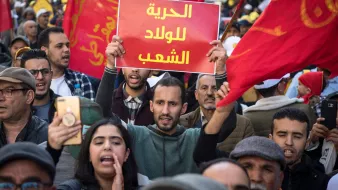 L'image montre une foule de manifestants, brandissant des drapeaux rouges. Au centre, un homme tient une pancarte avec le texte en arabe, tandis que d'autres personnes autour de lui semblent engagées dans la manifestation. On peut ressentir une atmosphère de protestation avec des expressions de détermination sur les visages des participants.
