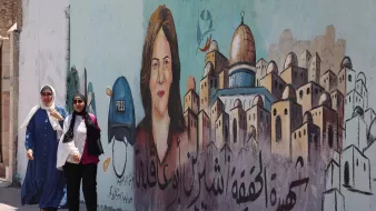 L'image montre deux femmes marchant le long d'un mur décoré d'une fresque. La fresque représente le portrait d'une femme avec un casque portant l'inscription "PRESS", suggérant qu'elle est journaliste. En arrière-plan, on aperçoit des maisons stylisées qui pourraient évoquer une ville ou un quartier particulier. Le texte en arabe est également visible, mais seul le contexte artistique et le message de solidarité ou de mémoire peuvent être interprétés. L'atmosphère est animée et engagée.