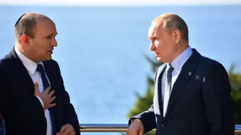 The image shows two men engaged in conversation by a seaside. One man is wearing a suit and has some form of head covering, indicating he may be of Jewish faith. The other man is also dressed in a suit and appears to be listening intently. They are both standing outdoors with a view of the water in the background. The setting suggests a formal or diplomatic meeting.