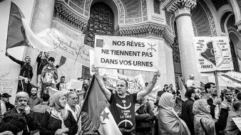L'immagine ritrae una manifestazione di protesta. Al centro, un uomo tiene in alto un cartello con scritto "NOS RÊVES NE TIENNENT PAS DANS VOS URNES", che significa "I nostri sogni non stanno nelle vostre urne". Sullo sfondo, ci sono altri manifestanti e striscioni recanti vari messaggi. L'atmosfera sembra intensa, con persone che esprimono il loro dissenso e la loro determinazione. L'immagine è in bianco e nero, aggiungendo un senso di drammaticità e urgenza alla scena.
