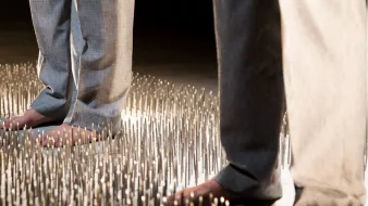 L'image montre deux personnes se tenant debout sur une surface couverte de nombreux picots métalliques. Les individus portent des pantalons clairs, et leurs pieds nus touchent les picots. La scène semble mettre en avant un contraste fort entre le confort des vêtements et l'inconfort potentiel de la surface sur laquelle ils se tiennent, créant une atmosphère à la fois intrigante et dérangeante.