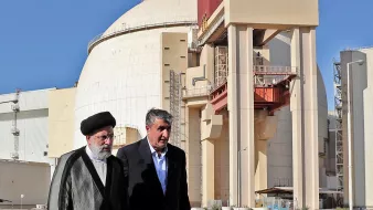 L'image montre deux hommes marchant devant une grande structure industrielle, qui semble être un réacteur nucléaire. Le bâtiment a une forme arrondie et est de couleur claire, avec des éléments de construction en béton visibles. Les deux hommes portent des vêtements formels ; l'un est vêtu d'un costume traditionnel et l'autre porte une veste. L'arrière-plan est dégagé, avec un ciel bleu. Cette scène pourrait illustrer une visite officielle ou une discussion liée à l'énergie nucléaire.